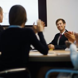 Group of business people meeting in corporate conference room, applauding at a coworker during his presentation. The man is showing charts and slides on a big TV monitor